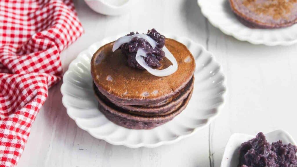 A stack of three purple pancakes topped with coconut strips and a dark paste, served on a white scalloped plate next to a red checkered cloth.