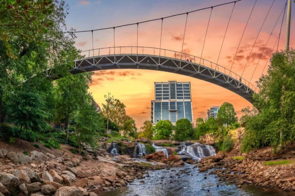 A pedestrian suspension bridge spans over a rocky stream and small waterfall in Greenville, South Carolina, with trees and modern buildings in the background at sunset.