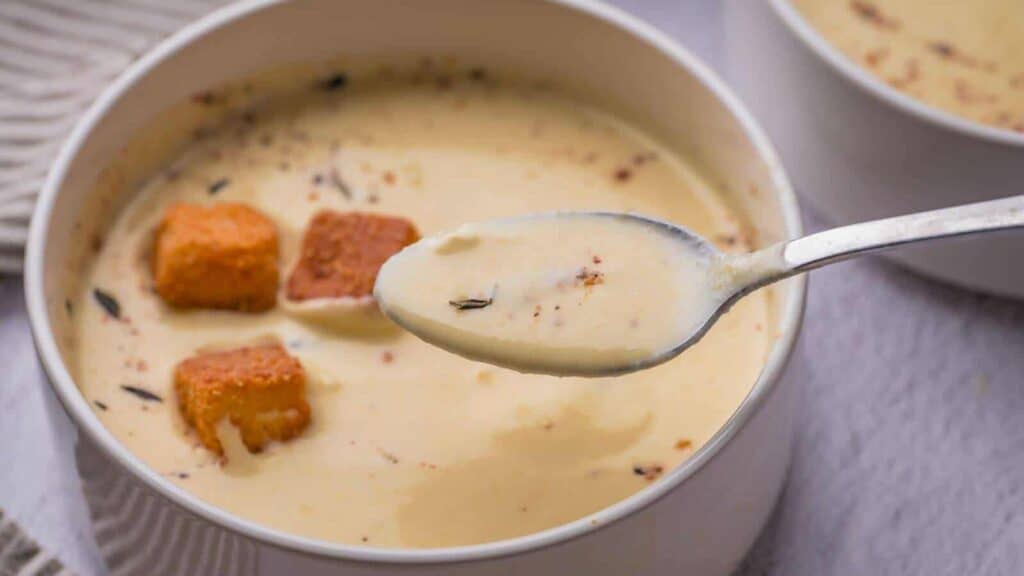 A close-up of a bowl of creamy soup with croutons and herbs, with a spoonful of soup held above the bowl.