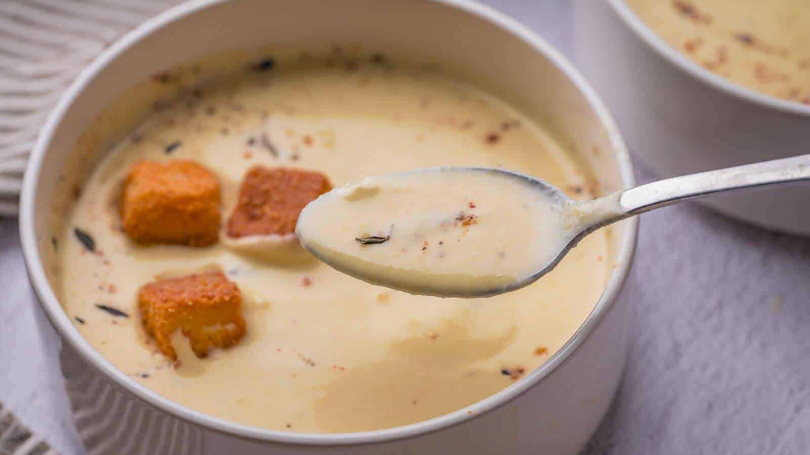 A bowl of creamy yellow squash soup with a few croutons, with a spoonful of soup held above the bowl.