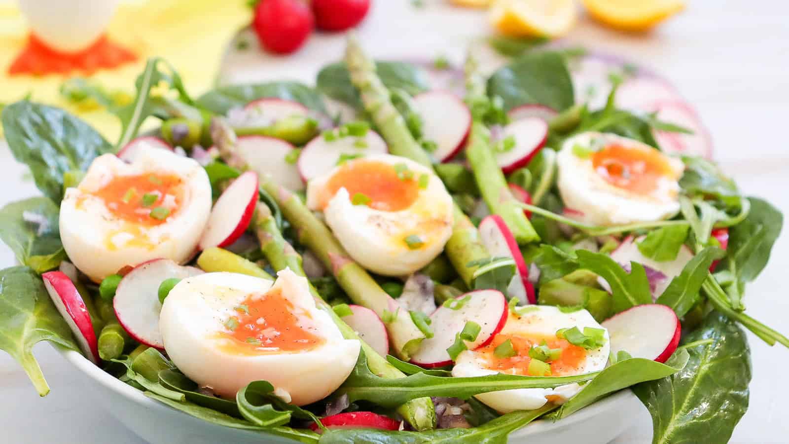 A vibrant asparagus salad with spinach, arugula, radishes, and soft-boiled eggs is topped with chopped green onions and arranged beautifully in a white bowl.