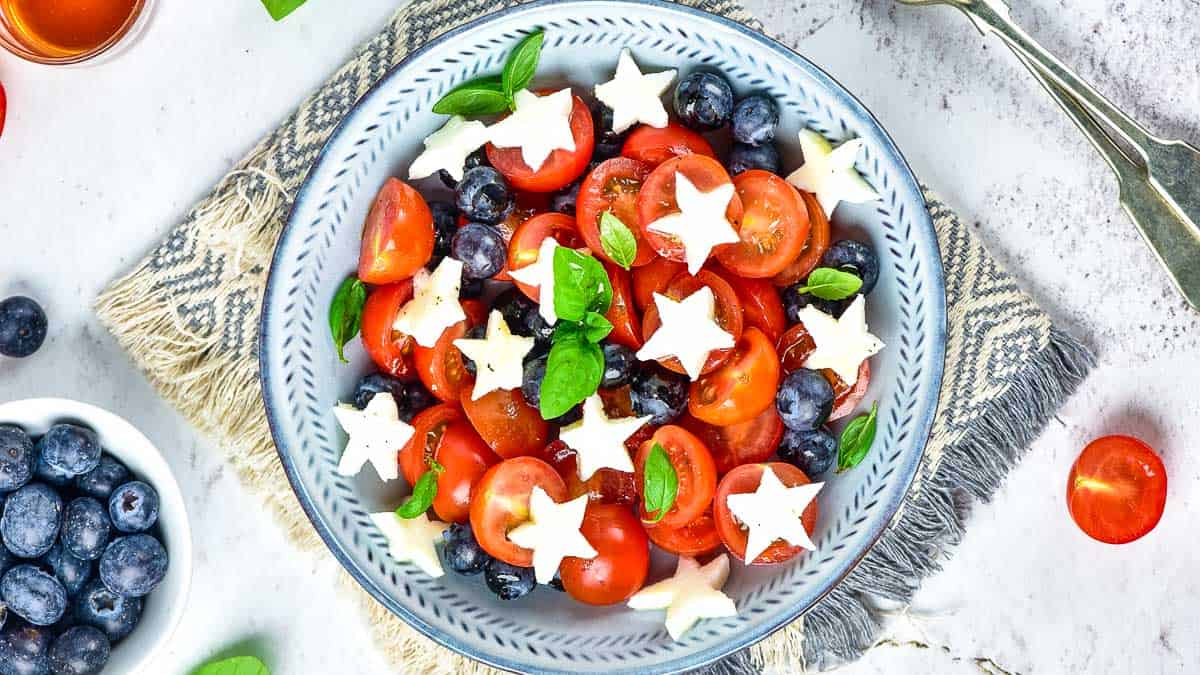 A bowl of salad with cherry tomatoes, blueberries, basil leaves, and star-shaped mozzarella pieces on a textured placemat.