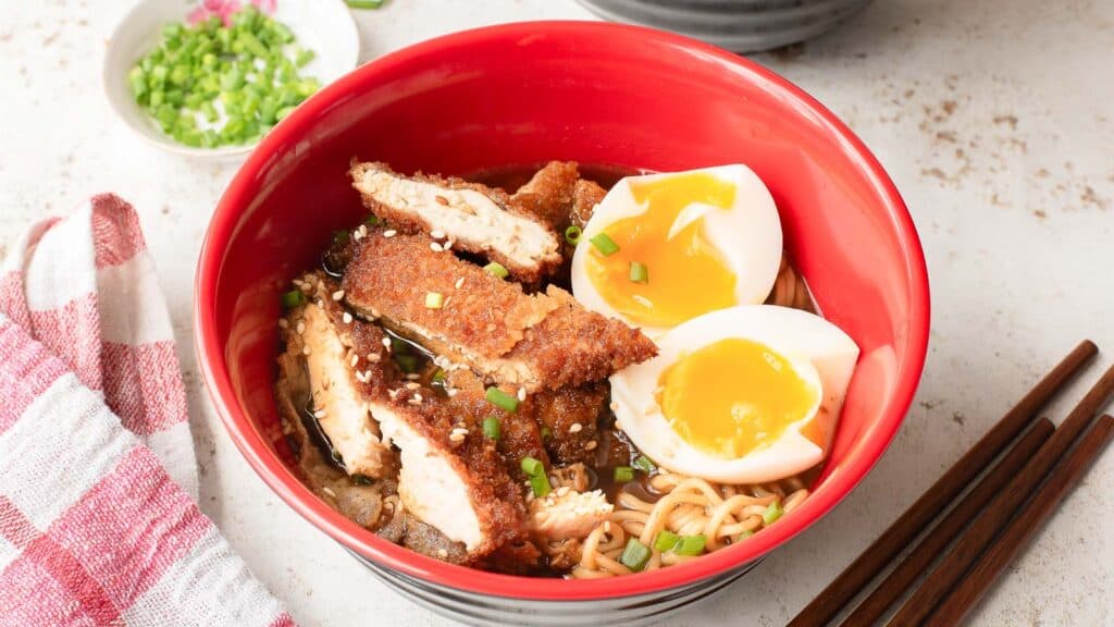 A bowl of ramen topped with sliced fried chicken, two soft-boiled eggs, chopped green onions, and sesame seeds, with chopsticks placed beside the bowl.