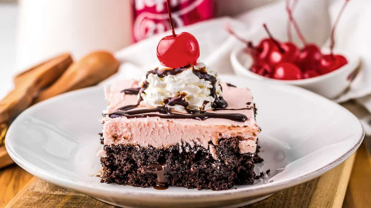 A slice of chocolate and pink cake topped with whipped cream, chocolate drizzle, and a cherry sits on a white plate, with more cherries in a bowl in the background.