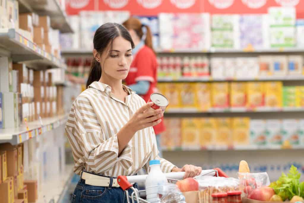 A woman stands in a grocery store aisle, examining a can while pushing a shopping cart filled with groceries.