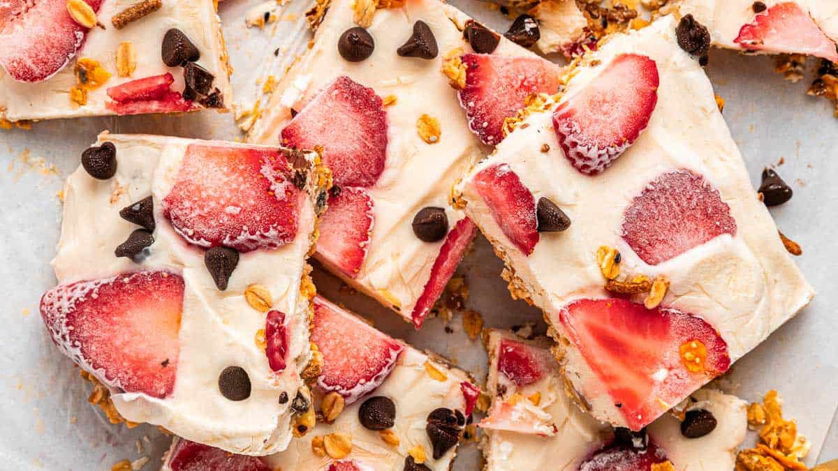 Squares of frozen yogurt bark topped with sliced strawberries, chocolate chips, and granola are arranged on a white surface.