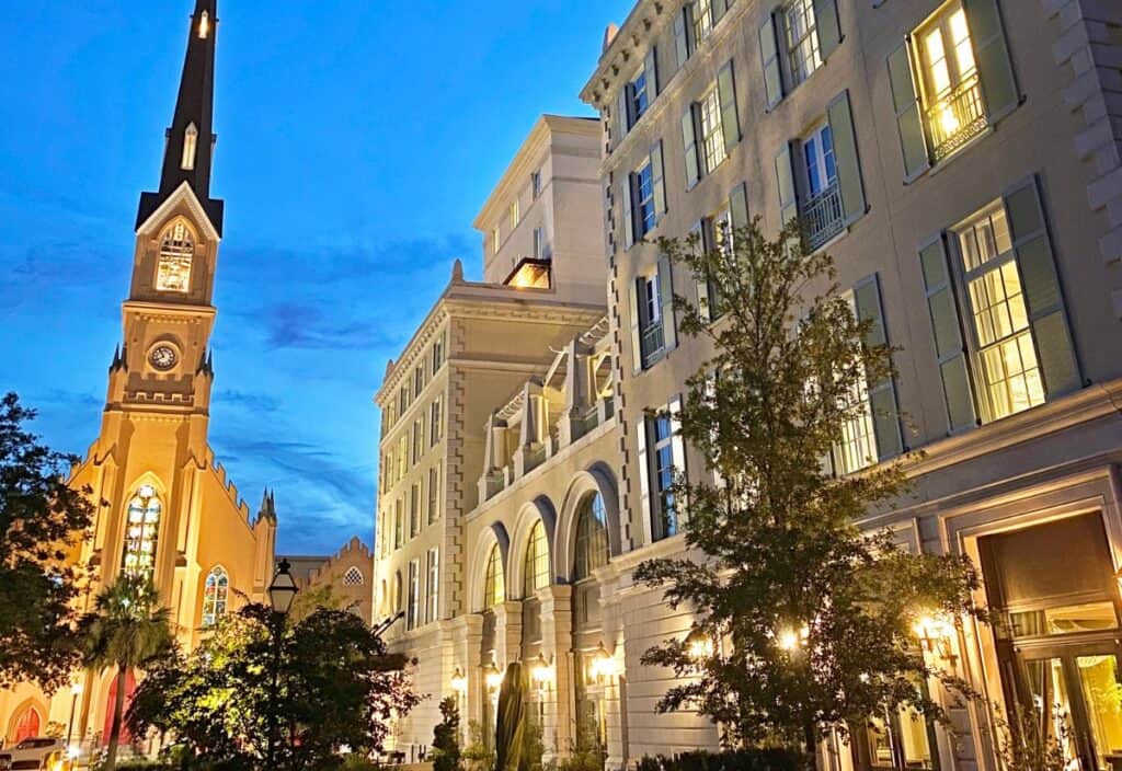 A church with a tall steeple stands next to one of the elegant Charleston hotels, its multi-story facade brightly lit at dusk, with trees in the foreground.