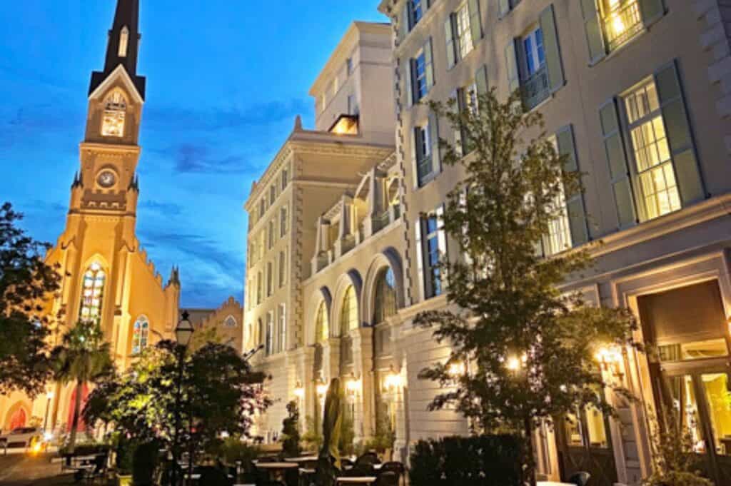 Evening view of a city street with a lit historic church on the left and a multi-story building with illuminated windows—possibly one of the charming Charleston hotels—on the right. Trees and an outdoor seating area are visible in the foreground.
