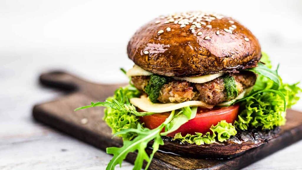 A hamburger with lettuce, tomato, cheese, and sauce, served on a large portobello mushroom cap instead of a bun, placed on a wooden board.