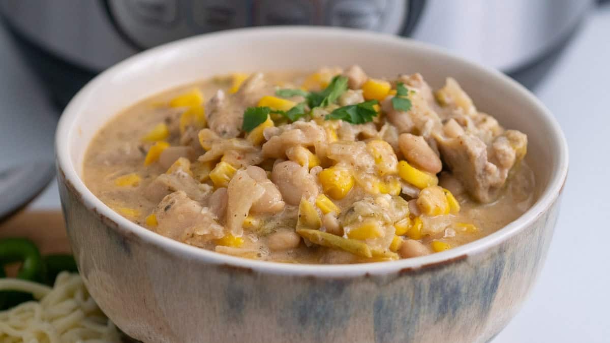 A bowl of white chili with chicken, white beans, corn, and green garnish, with a pressure cooker in the background.