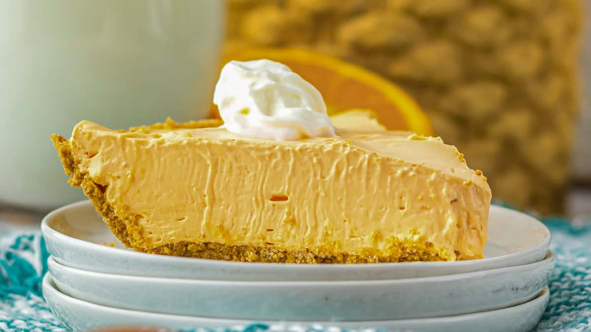 A slice of creamy orange pie with a graham cracker crust, topped with a dollop of whipped cream, served on stacked white plates.