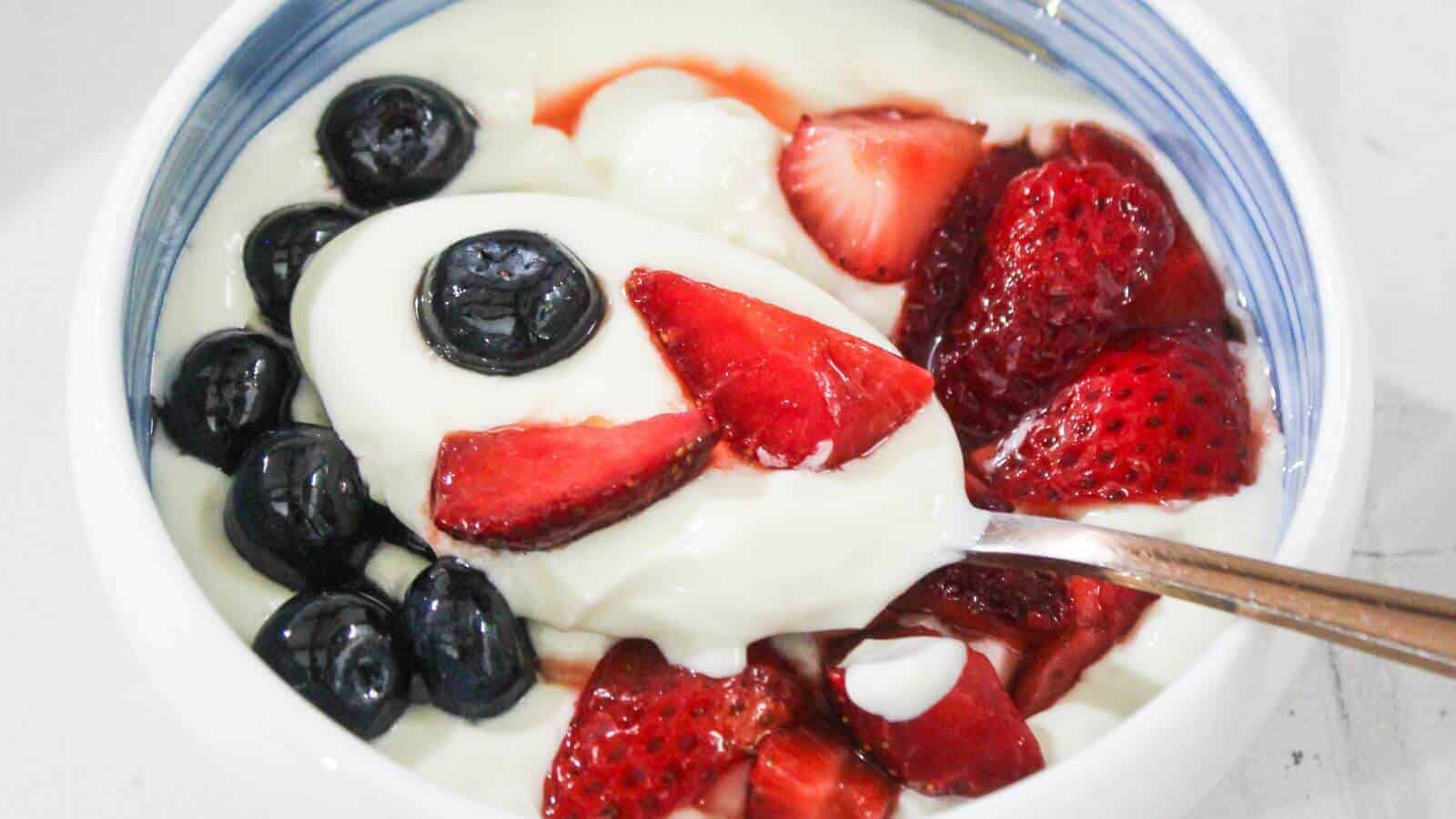 A bowl of whipped cream topped with blueberries and sliced strawberries, with a spoon resting inside.