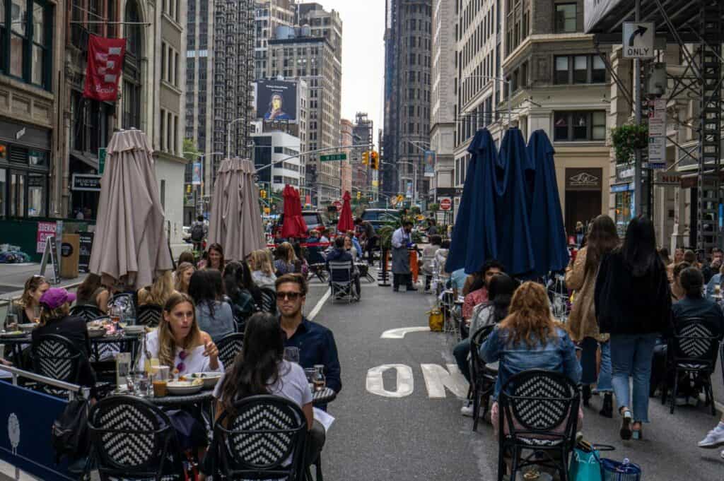 People dine at outdoor tables set up on a city street lined with tall buildings—a classic scene in one of America’s food cities, with umbrellas and traffic signs adding to the lively urban atmosphere.
