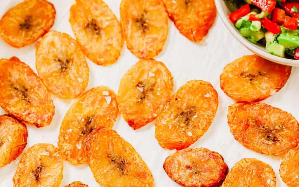 Golden-brown baked plantain chips are arranged on a white surface next to a bowl of diced vegetables.