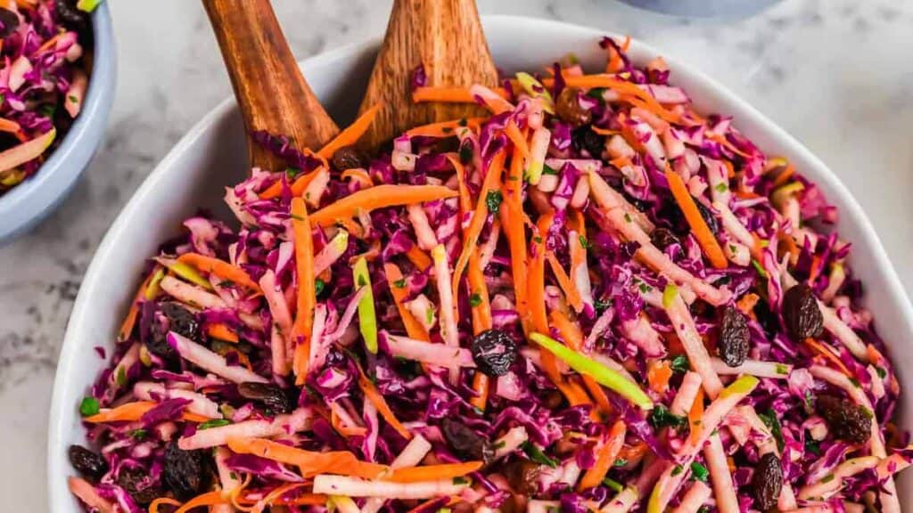 A bowl of colorful coleslaw with shredded red cabbage, carrots, green apple, and raisins, mixed with wooden salad servers.