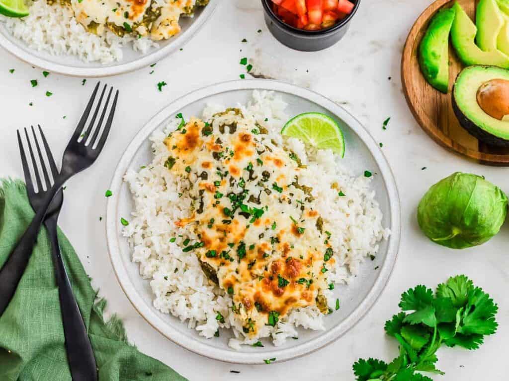A plate of white rice topped with cheesy baked chicken, garnished with chopped herbs and a lime wedge, served with sides of avocado, salsa, cilantro, and two black forks.