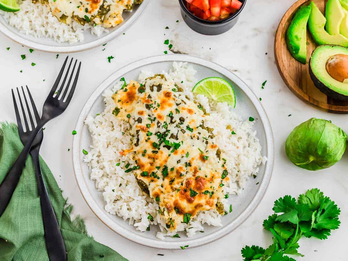 A plate of white rice topped with cheesy chicken and green salsa sauce with herbs, garnished with a lime.