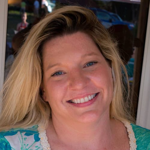 A woman with long blonde hair smiles at the camera, standing in front of a window with blurred people and outdoor scenery in the background.