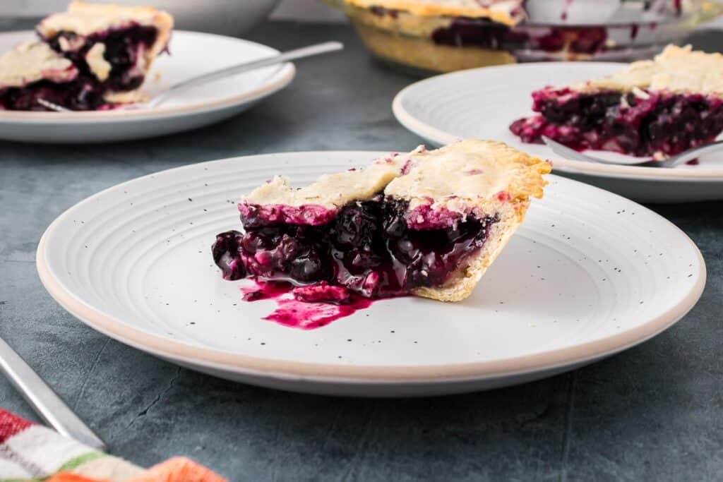 A slice of blueberry pie with a golden crust and visible berry filling sits on a white plate; additional pie slices are in the background.