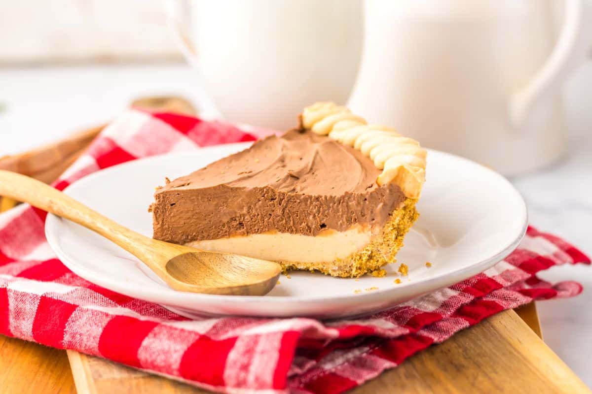 A slice of layered chocolate and peanut butter pie on a white plate with a wooden spoon, placed on a red and white checkered napkin.