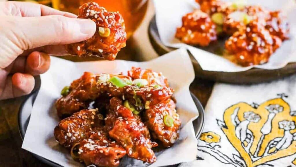 A hand holds a piece of saucy fried chicken over a plate filled with similar pieces, garnished with sesame seeds and green onions.