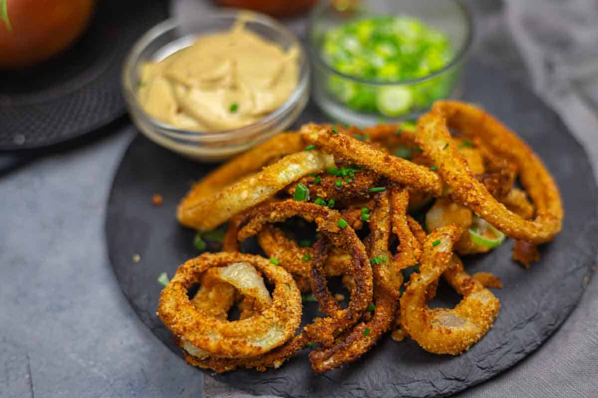 A serving of crispy fried onion rings on a dark plate, garnished with chopped herbs, with dipping sauce and chopped green onions in small glass bowls.
