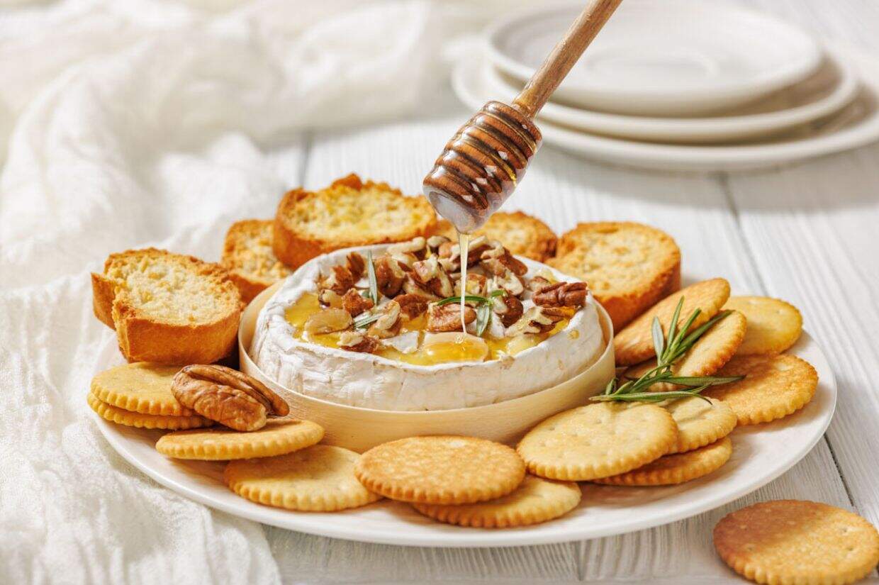 A plate with Baked Brie with Honey and Pecans  surrounded by crackers and bread slices, with a honey dipper drizzling honey over the cheese.