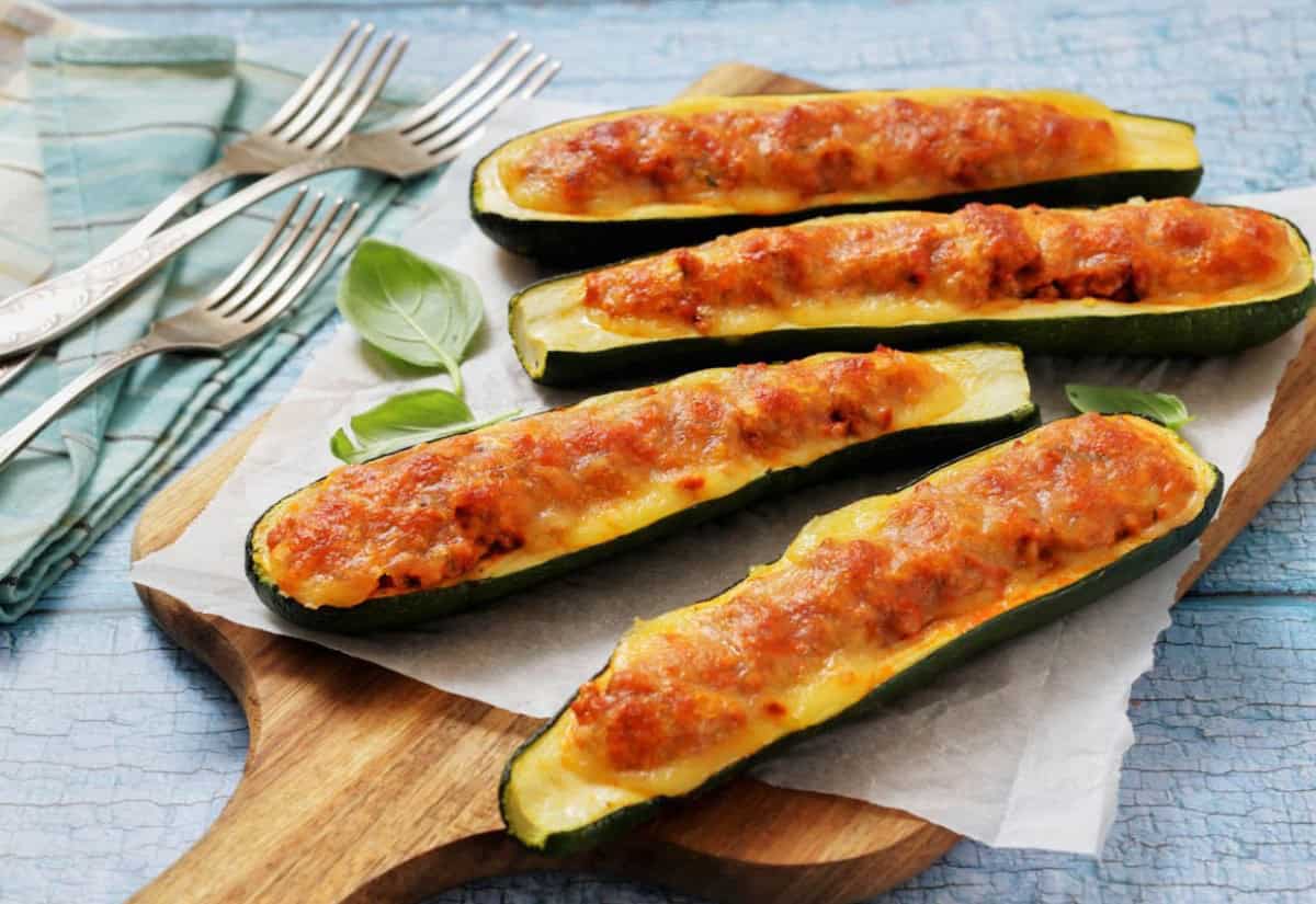 Baked zucchini boats stuffed with a rich meat and cheese topping on a wooden board. Four silver forks rest on a blue patterned cloth nearby.