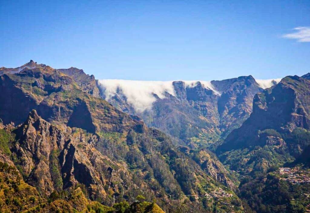 Mountain range on Madeira Island with steep rocky cliffs, lush green vegetation, and a layer of clouds spilling over the ridge under a clear blue sky.
