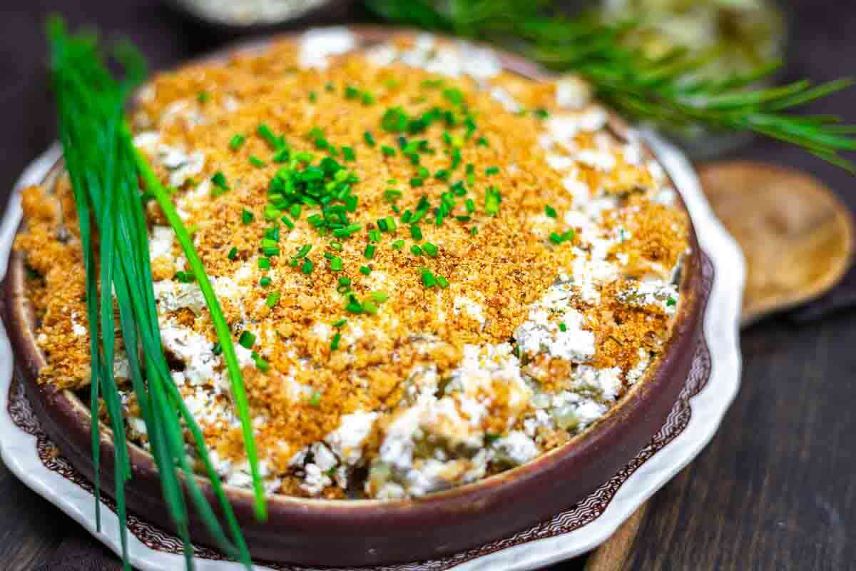A baked dill pickle dip topped with golden breadcrumbs and chopped chives, garnished with fresh herbs on the side, served in a round dish.
