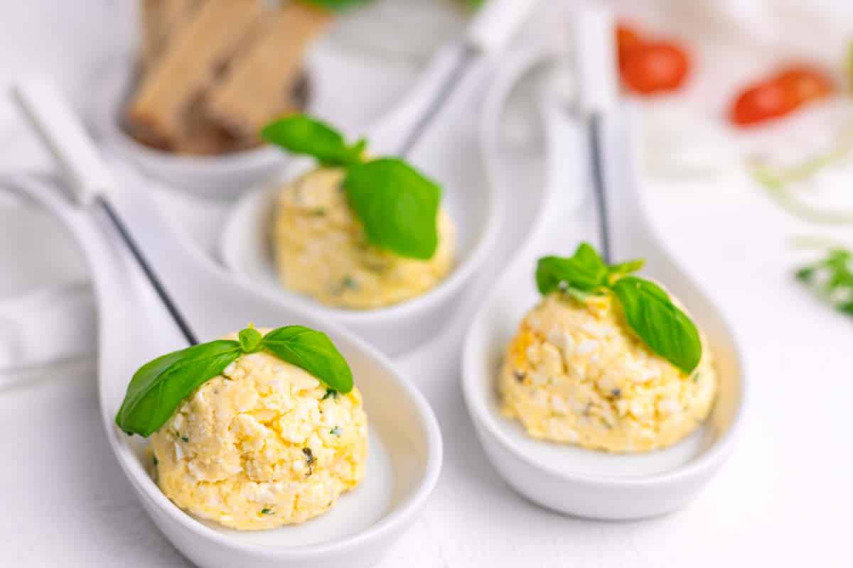 Three white spoons with scoops of cheese topped with basil leaves, placed on a white surface.