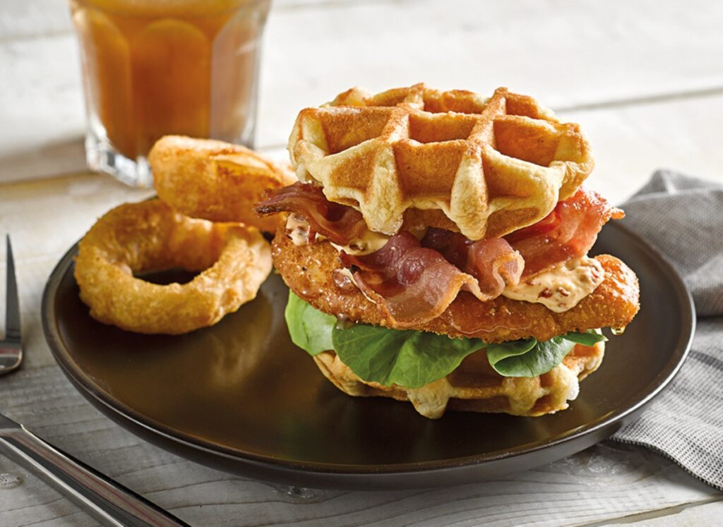 A sandwich with waffles as bread, filled with fried chicken, bacon, lettuce, and sauce, is served on a dark plate with onion rings and a drink in the background&mdash;a creative twist often rivaled by brioche in kitchens and cafes.