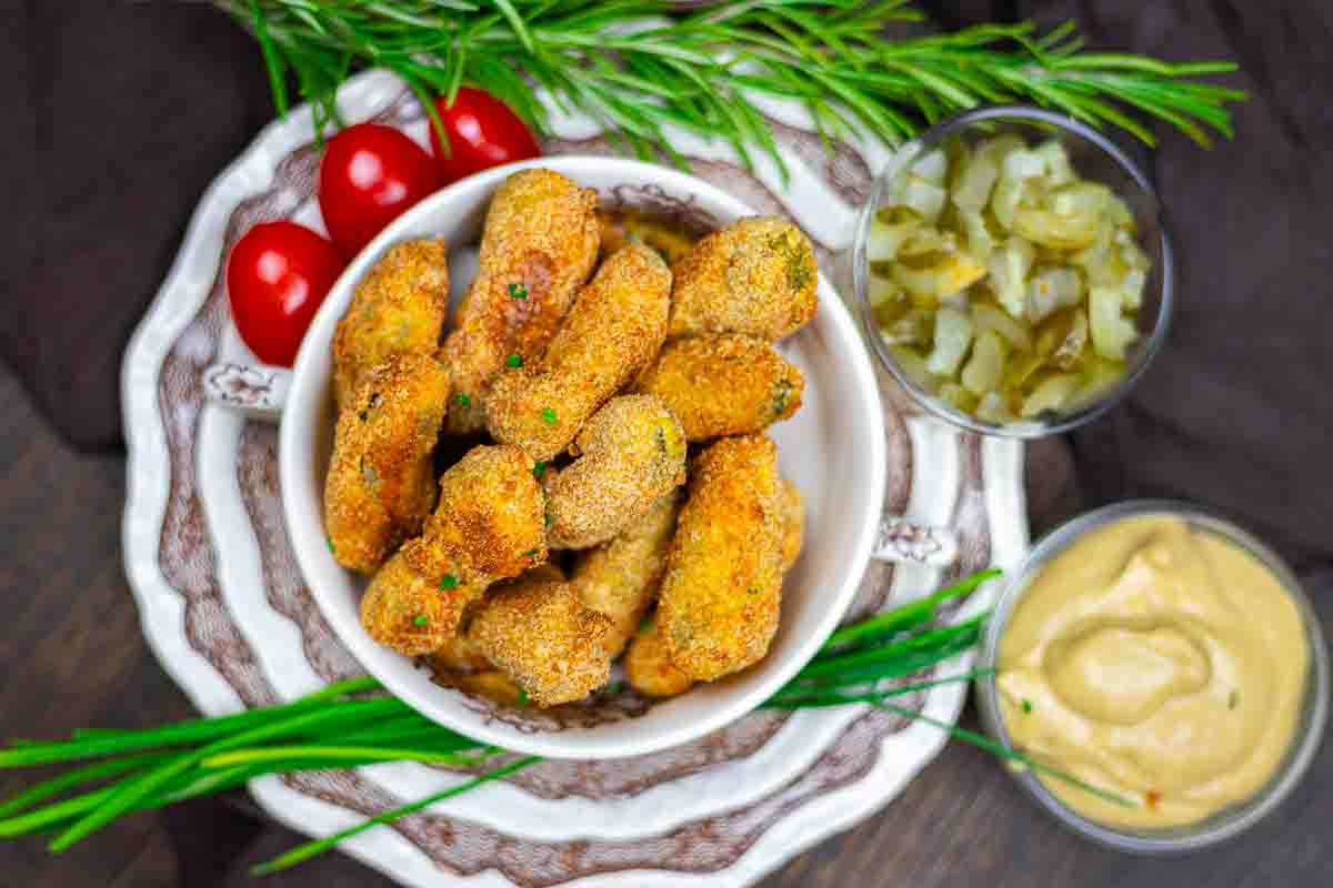 A bowl of breaded, fried dill pickles on a plate, served with sliced pickles, sauce, cherry tomatoes, and fresh herbs.