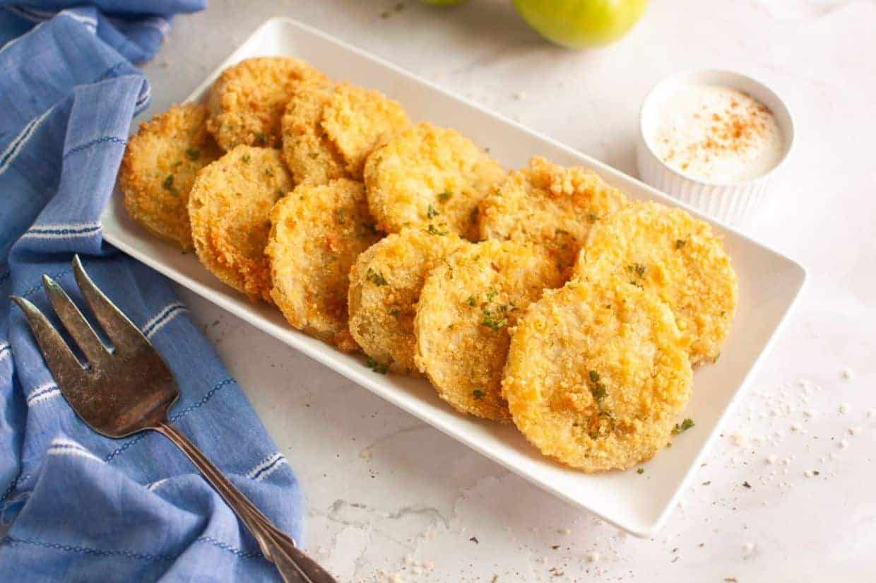 A rectangular white plate with fried green tomato slices garnished with herbs, a small cup of dipping sauce, a fork, and a blue napkin on the side.