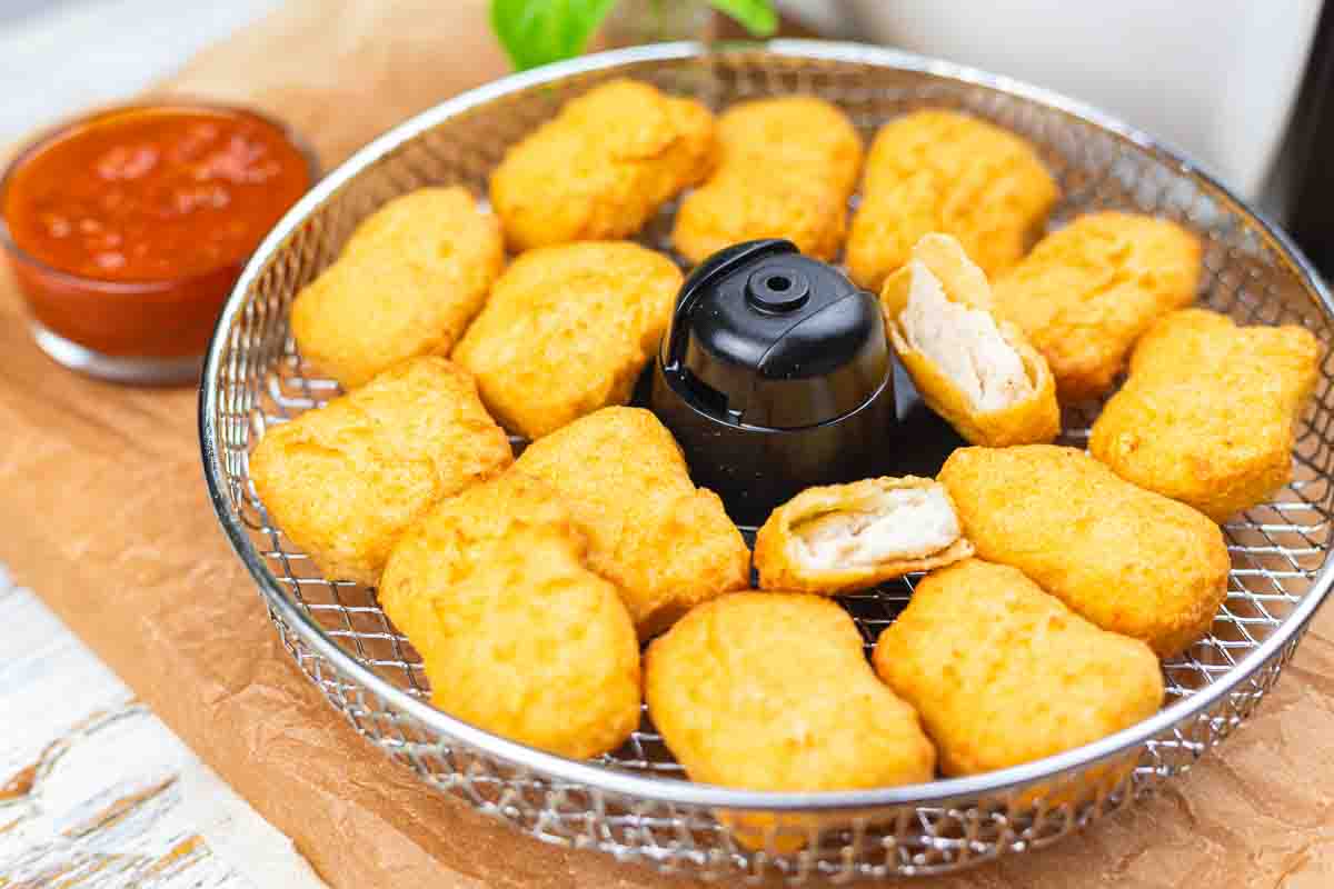 A wire basket filled with golden-brown chicken nuggets, some bitten, with a small cup of red dipping sauce beside it.