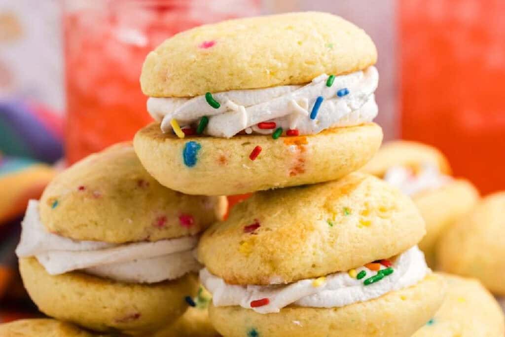 Three funfetti whoopie pies with white frosting and colorful sprinkles are stacked on a table, with a blurred pink drink in the background.