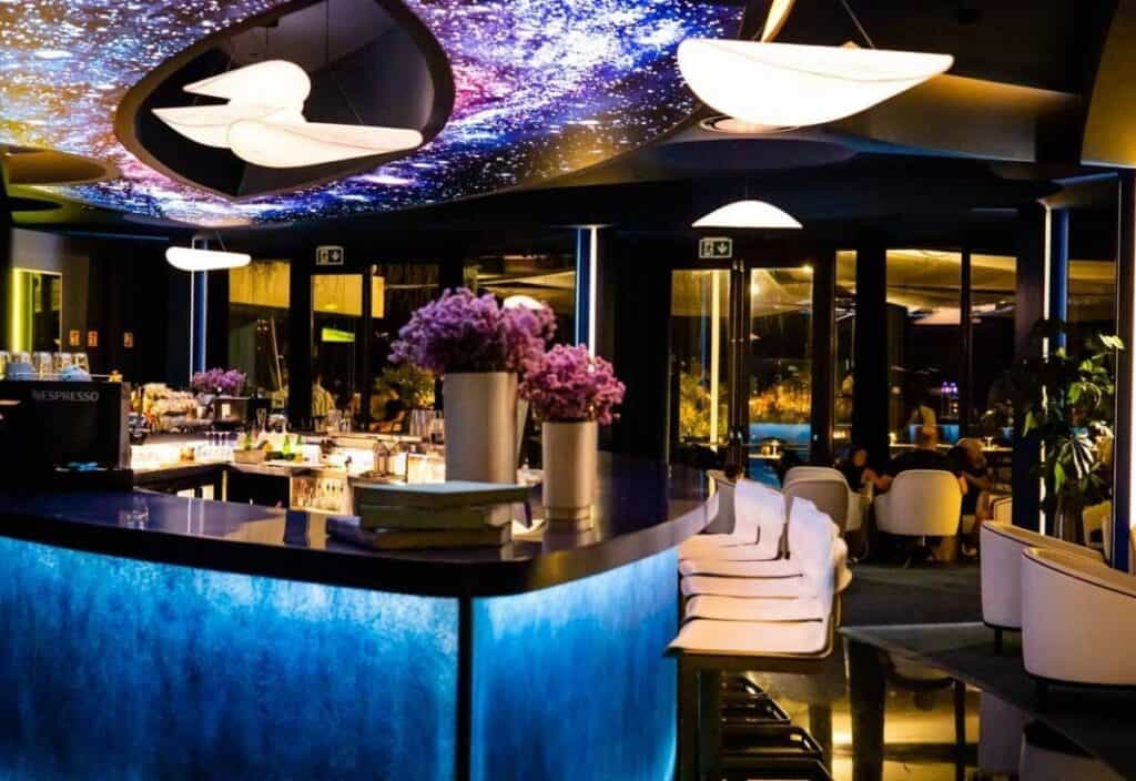 Modern bar on Madeira Island with a glowing blue counter, white chairs, flower arrangements, and a ceiling featuring galaxy-inspired lighting. Patrons are seated in the background.