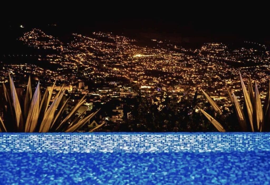A nighttime cityscape with numerous lights, viewed from behind a mosaic-tiled pool and lush plants in the foreground, captures the enchanting beauty of Madeira Island.