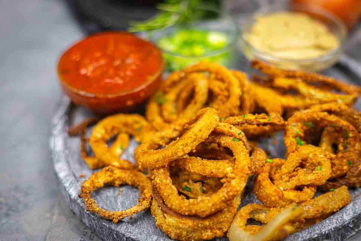A plate of crispy fried onion rings with a side of red dipping sauce and a bowl of mustard, garnished with green herbs.