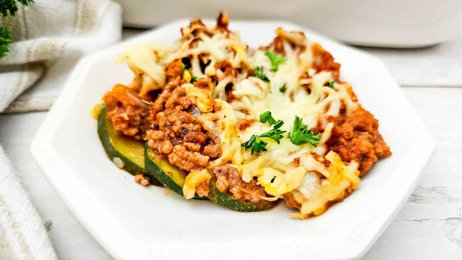 A serving of ground beef and zucchini bake topped with melted cheese and garnished with parsley is presented on a white plate.