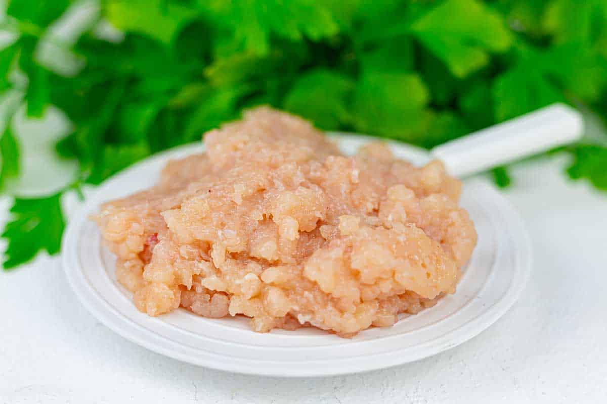 Plate of raw ground chicken on a white surface, with leafy greens in the background.