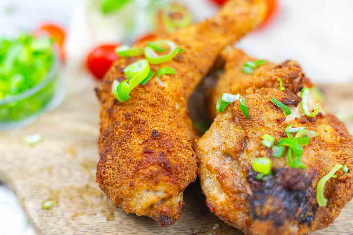 Two pieces of crispy fried chicken garnished with chopped green onions on a wooden surface, with a bowl of sliced green onions and tomatoes in the background.