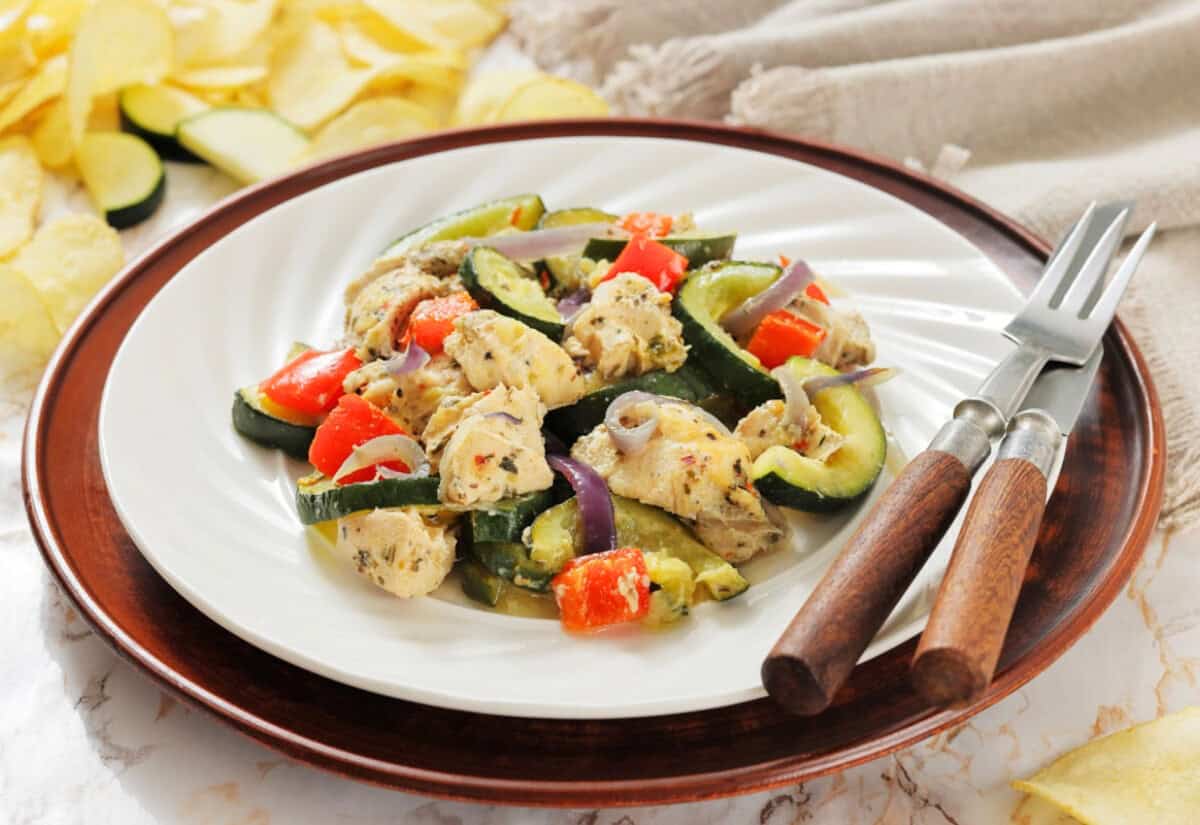 Plate of seasoned chicken, zucchini, red bell peppers, and red onions on a wooden-rimmed plate. Rustic backdrop with fork and knife. Cozy and appetizing.