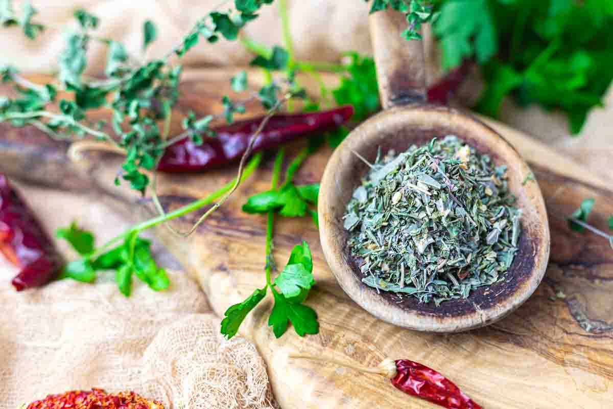 A wooden spoon filled with dried mixed herbs sits on a rustic board, surrounded by fresh parsley, dried chili peppers, and sprigs of fresh herbs.