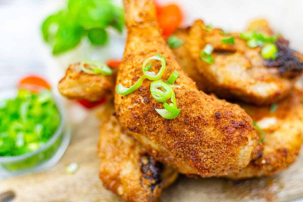 Close-up of crispy chicken legs garnished with sliced green onions, with a side of chopped herbs and vegetables in the background.