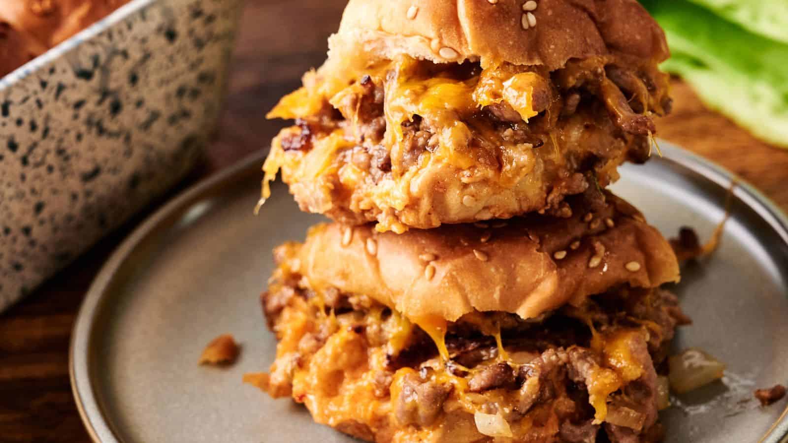 Two sloppy joe sandwiches with melted cheese on sesame seed buns are stacked on a gray plate.