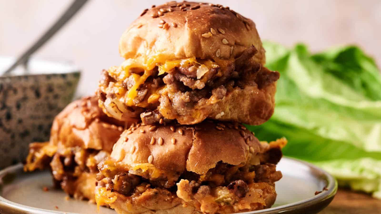 Two sloppy joe sliders with melted cheese and ground beef filling stacked on a plate, with lettuce and a bowl in the background.