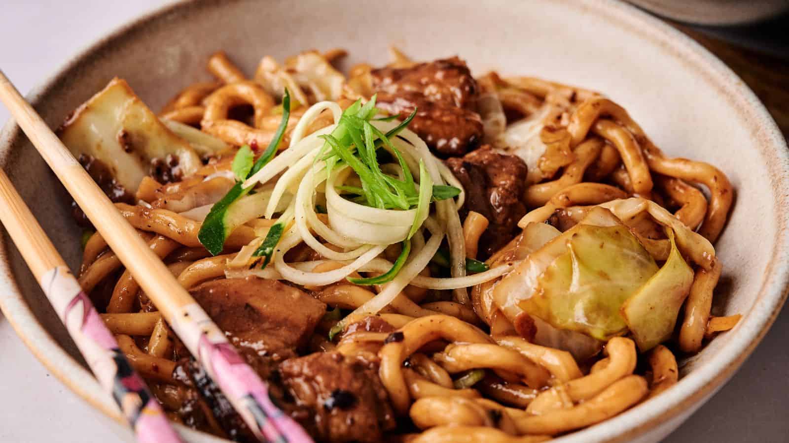 A bowl of stir-fried udon noodles with beef, cabbage, and green onion garnish, served with a pair of chopsticks on the side.