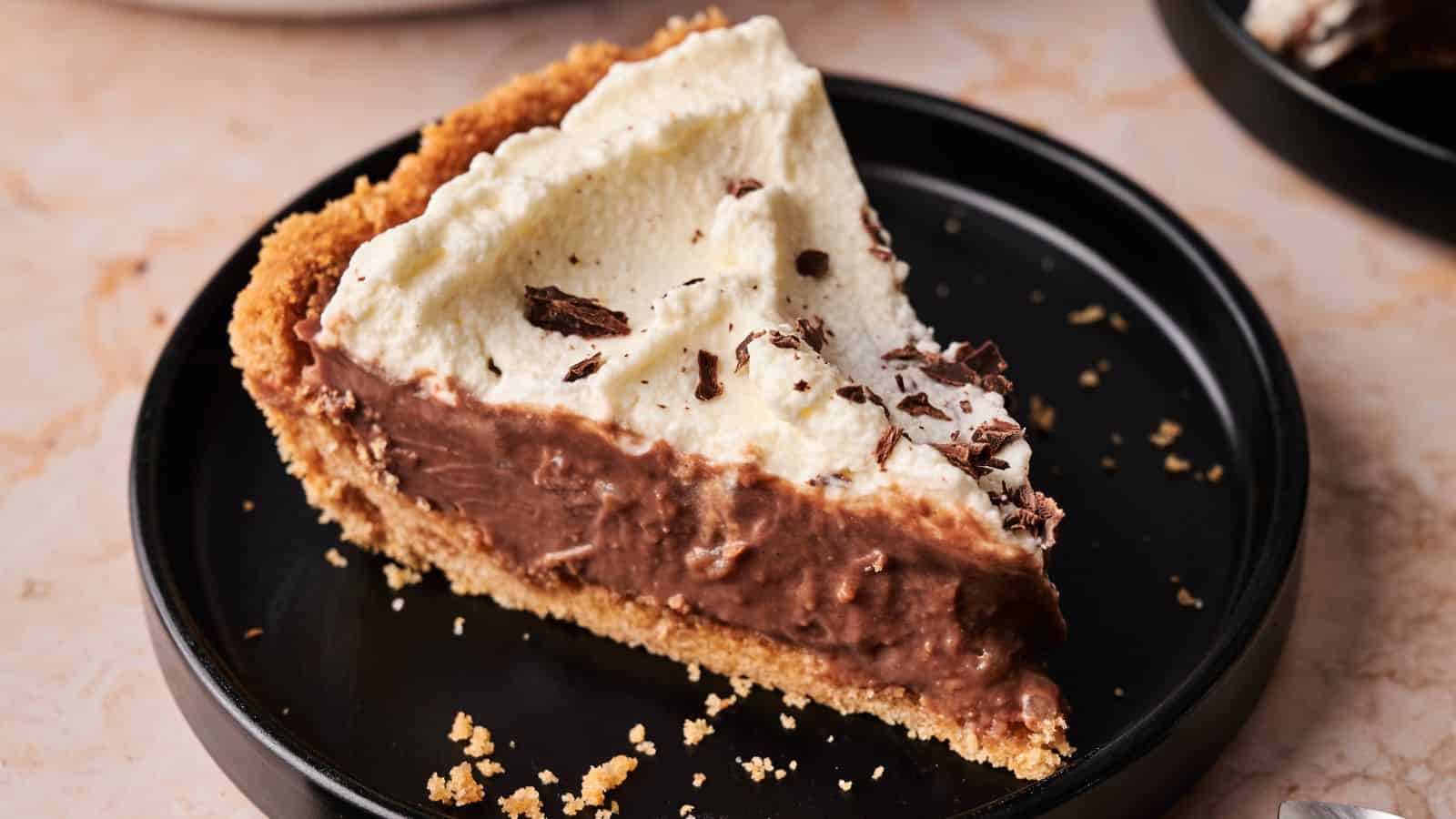 A slice of chocolate cream pie with a graham cracker crust, topped with whipped cream and chocolate shavings, served on a black plate.