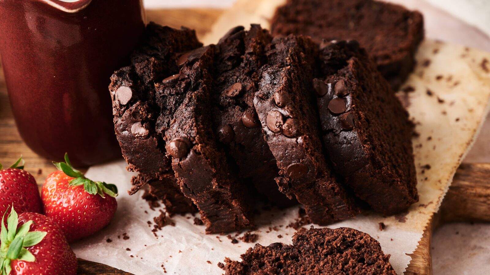 Sliced chocolate loaf cake with chocolate chips on parchment paper, next to a jar and fresh strawberries.